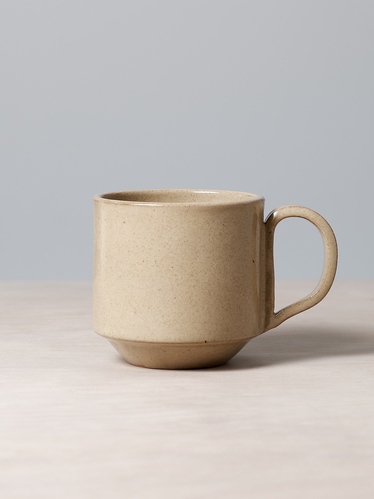 The Richard Beauchamp Tall Stacking Mug – Sand, featuring a curved handle and beige ceramic with a subtle sand glaze, is placed on a light wooden surface against a gray background.