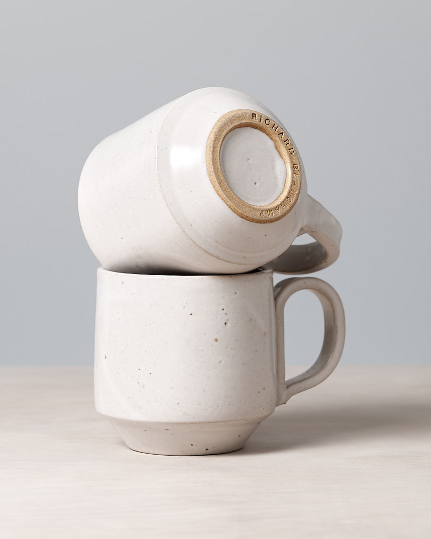 A Richard Beauchamp Tall Stacking Mug – White, rests elegantly on a wooden surface against a plain gray background, evoking the artisanal charm of Ōtautahi Christchurch.
