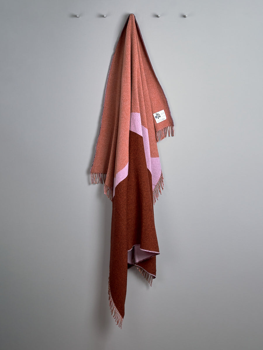 A Dune Blanket by Seljak Brand, brown and pink in color, is hanging on a wall.