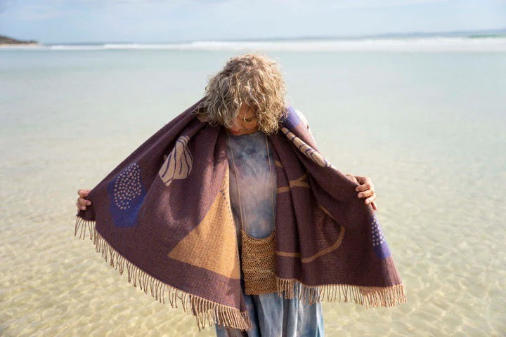 A woman standing on the beach holding a Seljak Brand Gather Blanket.