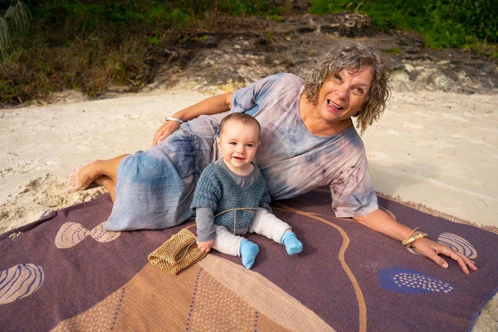 An older woman with a baby sitting on a Seljak Brand Gather Blanket on the beach.