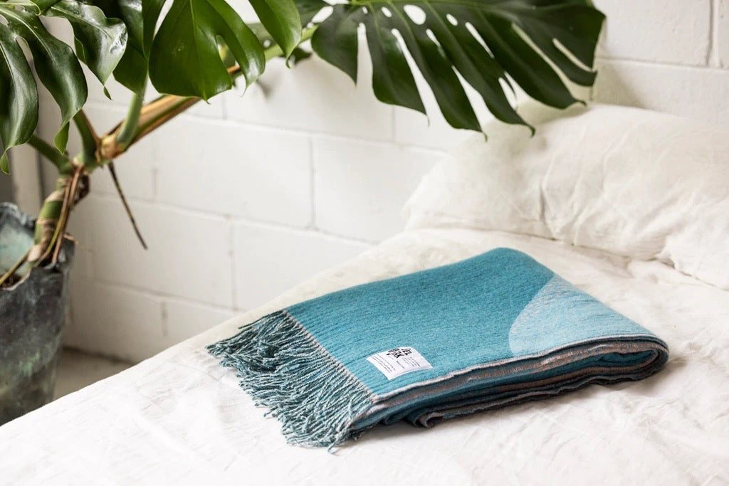 A Seljak Brand Passage Blanket on a bed with a plant in the background.