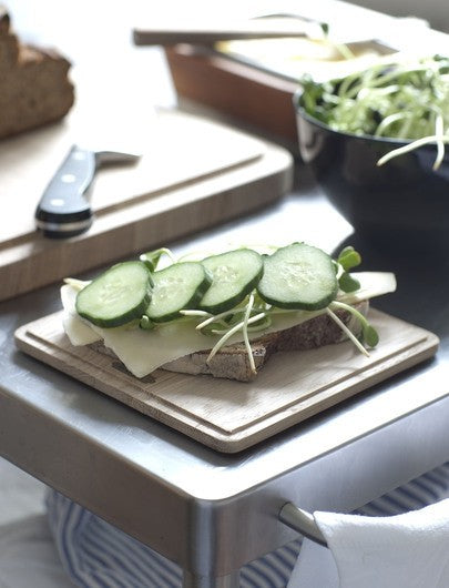 A Skagerak wooden cutting board, the Plank Square Trencher 4pcs – Oak, with a sandwich and cucumbers on it.