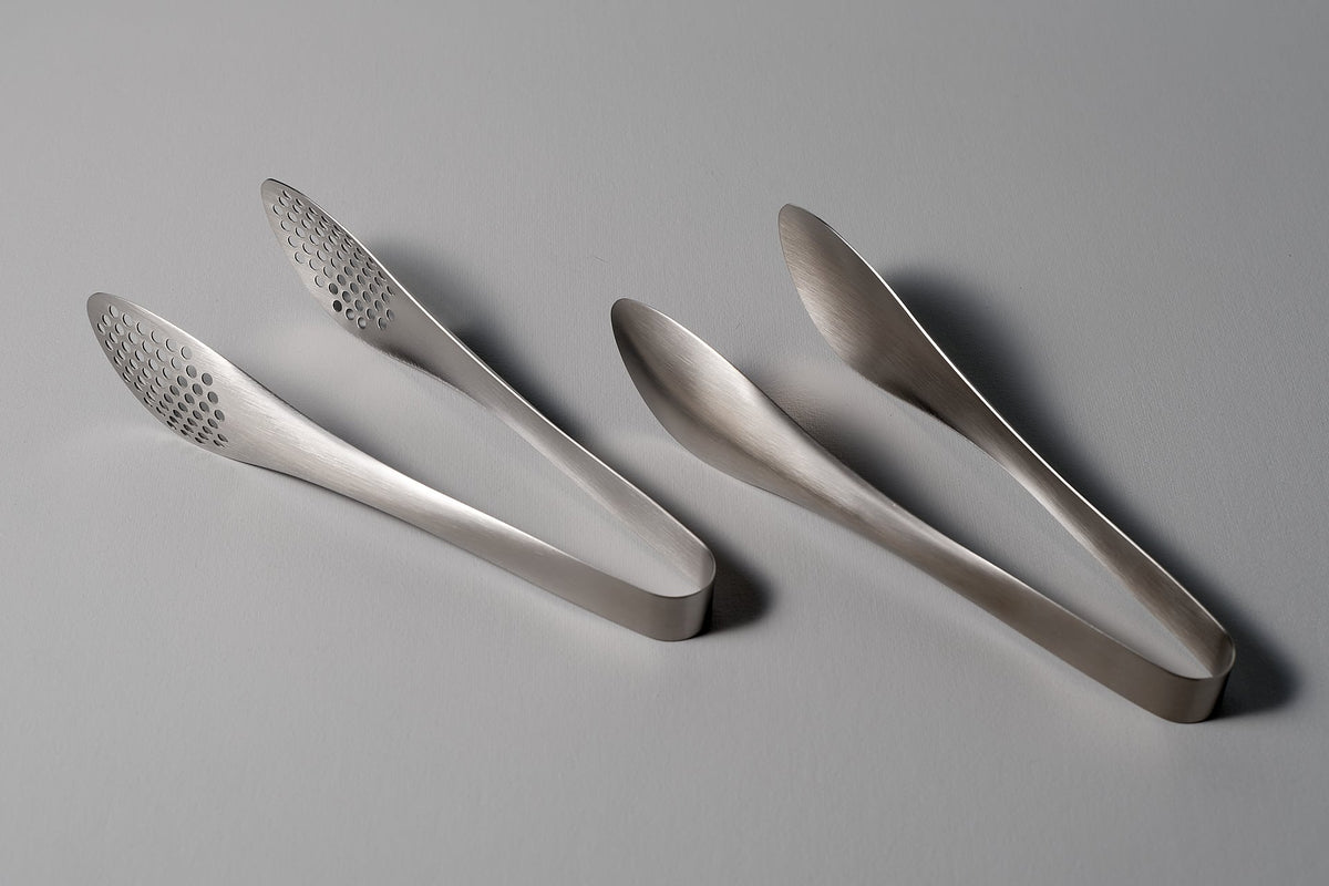 Two Sori Yanagi stainless steel tongs - perforated on a grey surface.