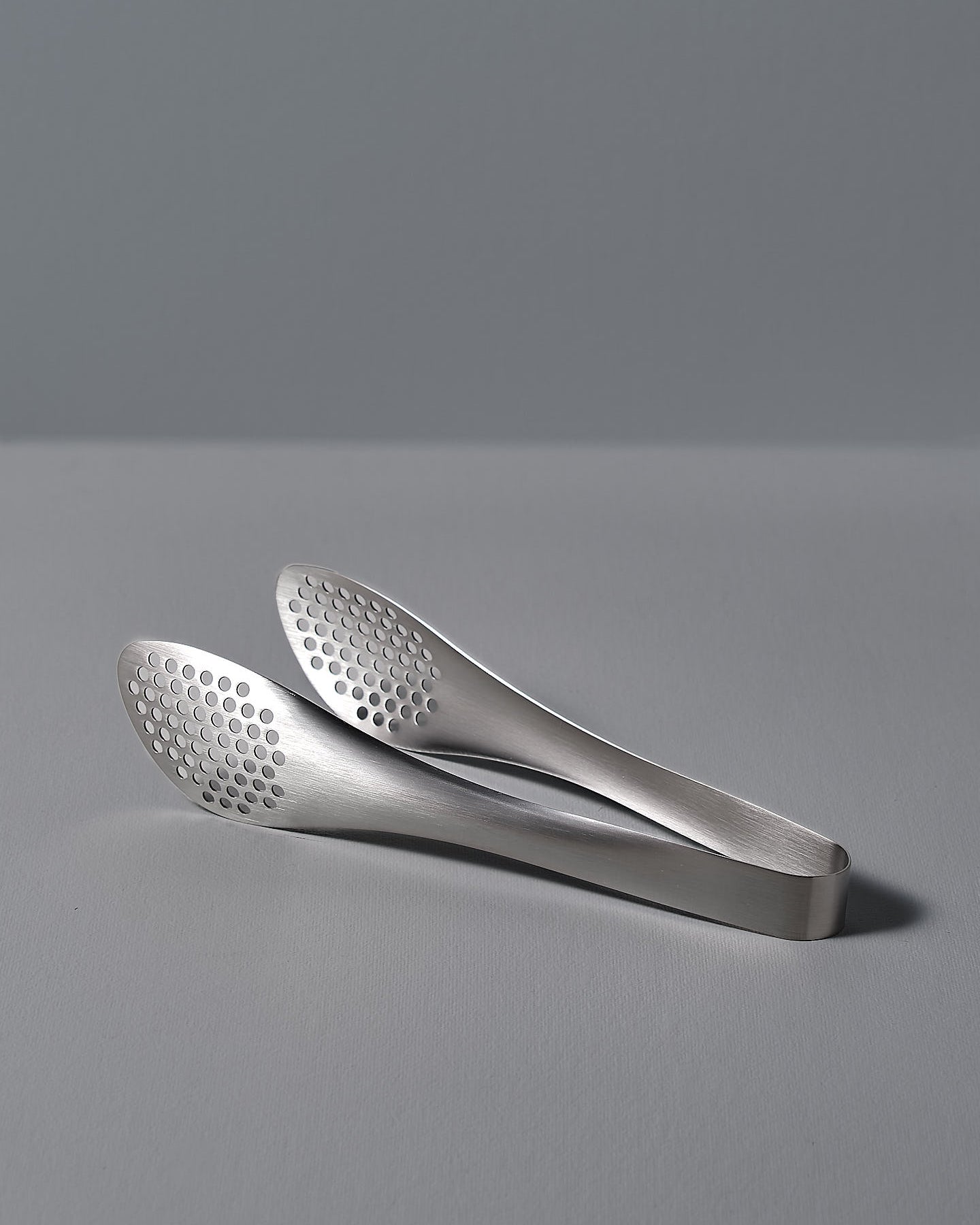 A pair of Sori Yanagi stainless steel tongs - perforated on a white surface.