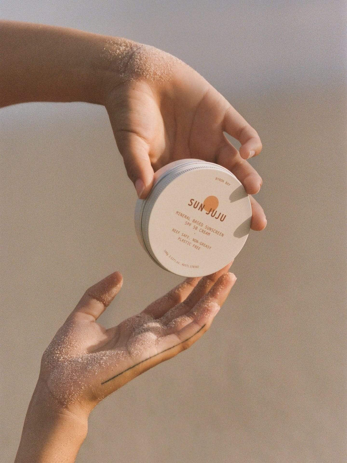 A person&#39;s hand holding a Sun Juju SPF50 Sunscreen for Face &amp; Body ⋅ Hydrating Reef Safe – 100𝚐 on the beach.