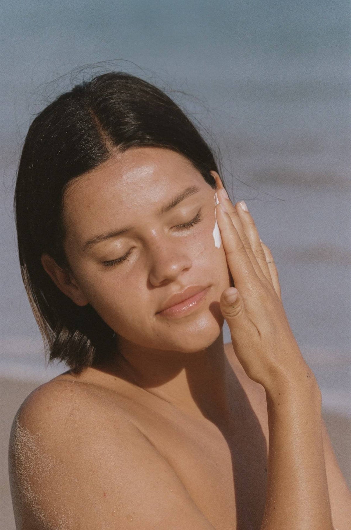 A woman applying Sun Juju SPF50 Sunscreen for Face &amp; Body ⋅ Hydrating Reef Safe – 100𝚐 to her face on the beach.