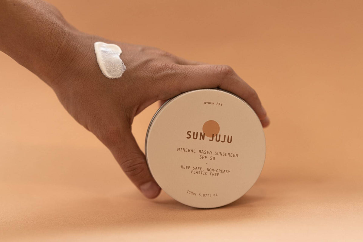 A hand is holding a Sun Juju SPF50 Sunscreen for Face &amp; Body ⋅ Hydrating Reef Safe – 100𝚐 on a beige background.