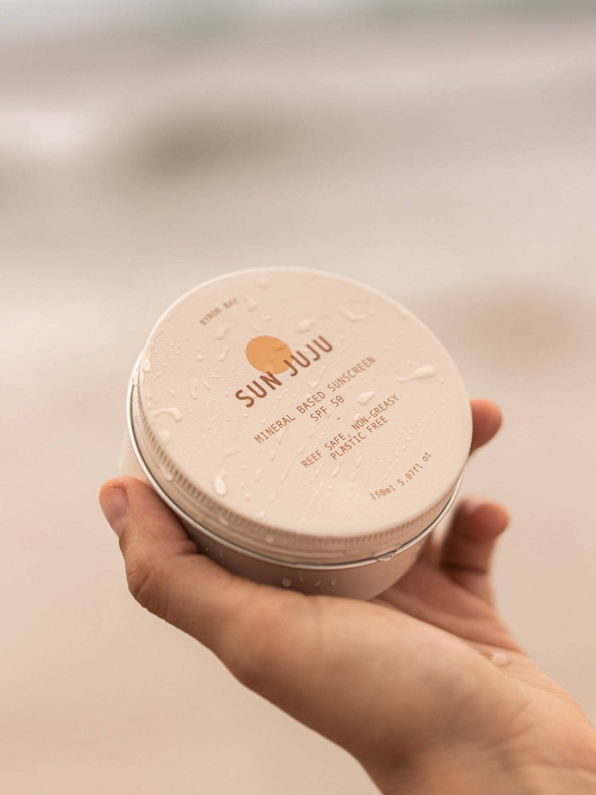 A person holding a tin of Sun Juju SPF50 Sunscreen for Face &amp; Body ⋅ Hydrating Reef Safe – 100𝚐 on the beach.