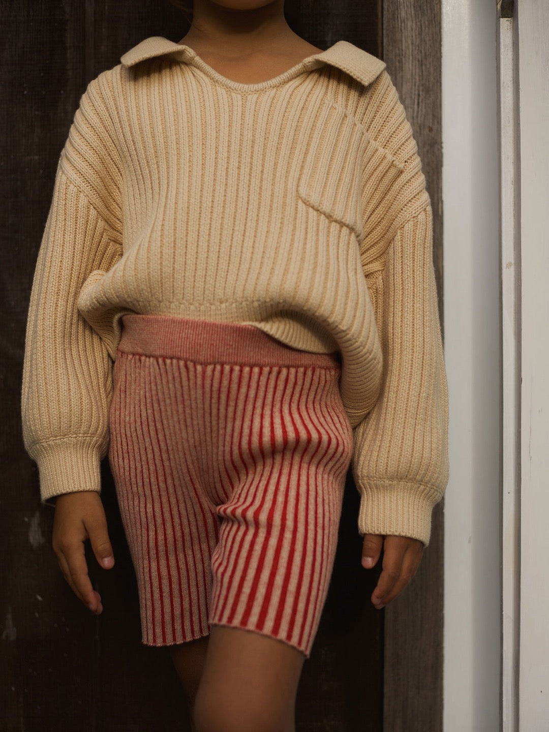 A young girl wearing a beige sweater and Rue Short – Red Multi striped shorts from SUNNA STUDIOS.