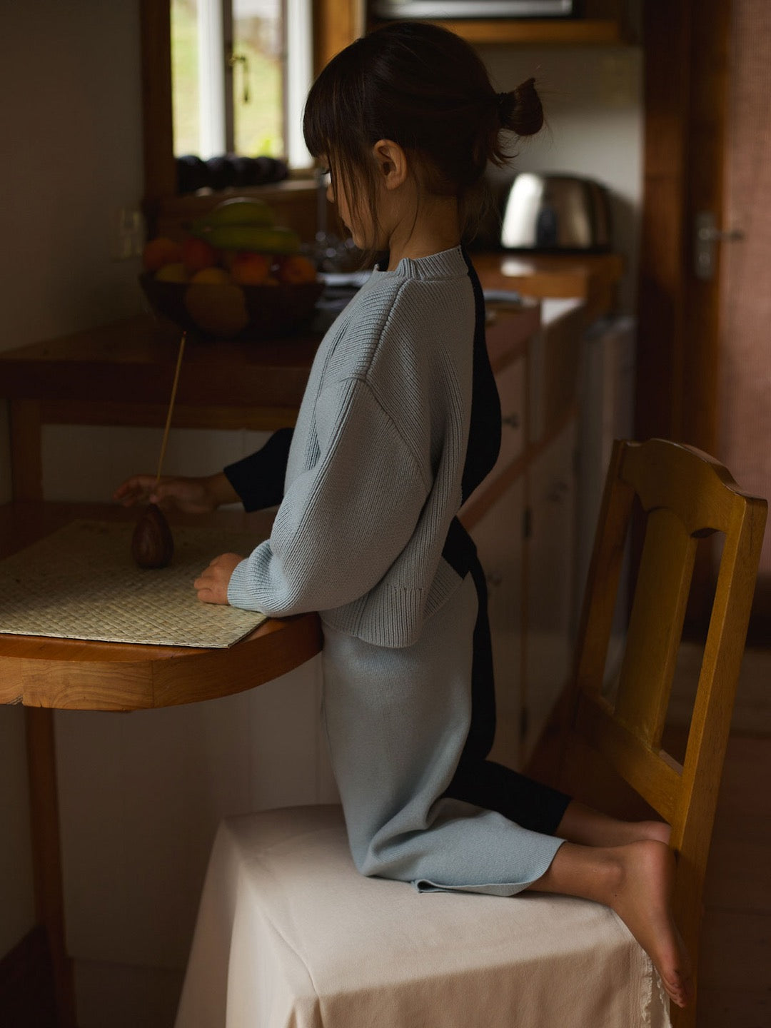 A little girl sitting on a chair in a kitchen wearing the SOL Sweater – Blue Multi by Sunna Studios.