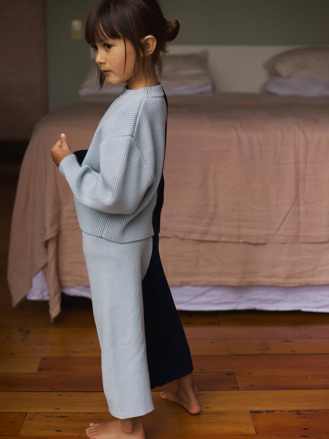 A little girl standing in a bedroom wearing a SUNNA STUDIOS Sol Sweater – Blue Multi.