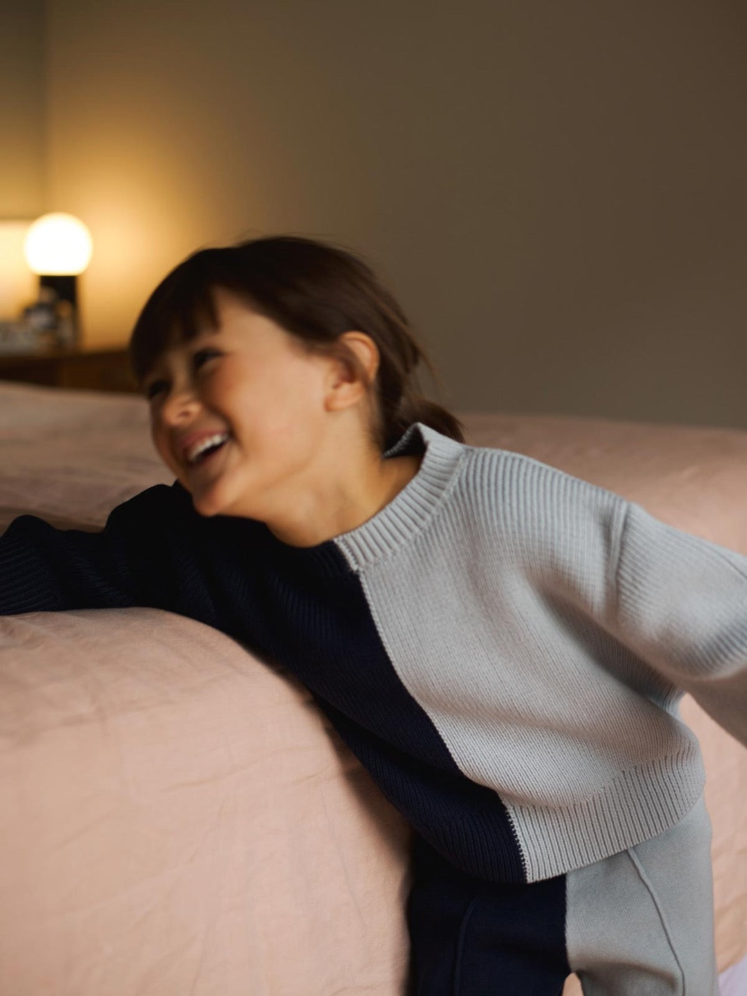A little girl is laughing on a bed while wearing the SUNNA STUDIOS Sol Sweater – Blue Multi.