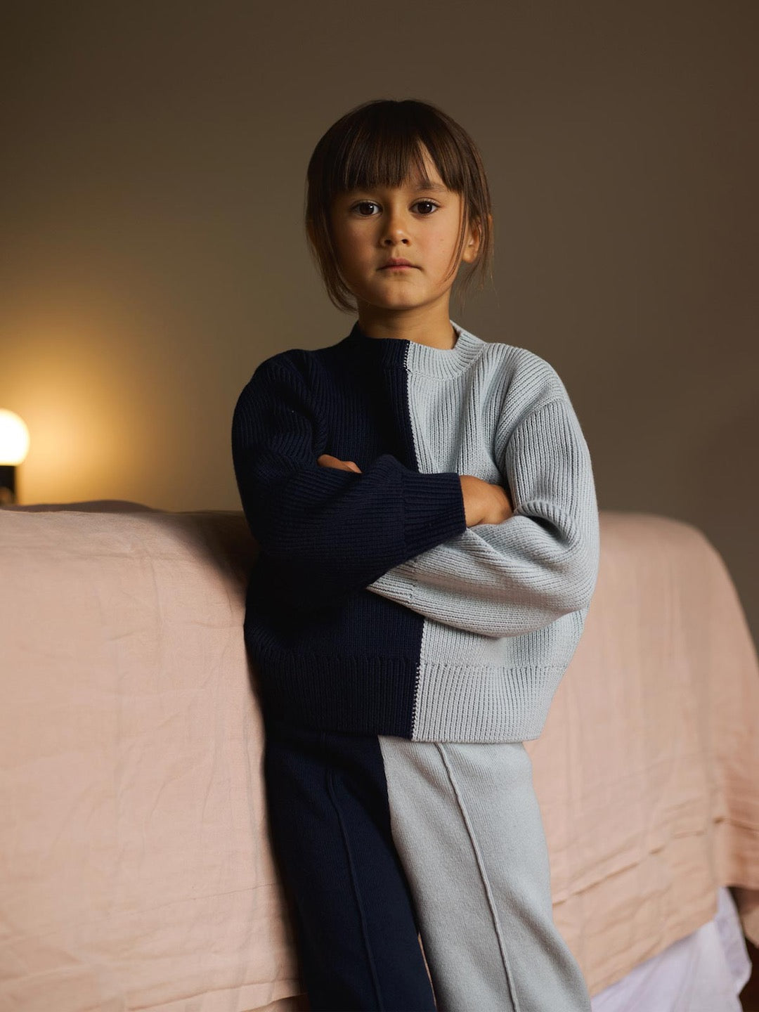 A young girl is standing on a bed with her arms crossed, wearing the Sol Sweater – Blue Multi by SUNNA STUDIOS.