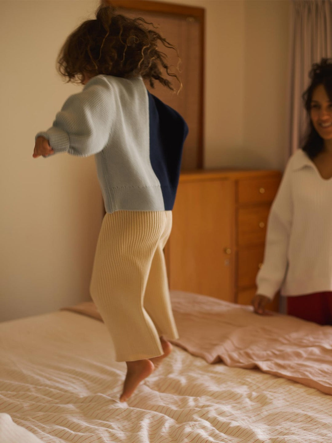 A little girl jumping on a bed wearing the Sol Sweater - Blue Multi by SUNNA STUDIOS.