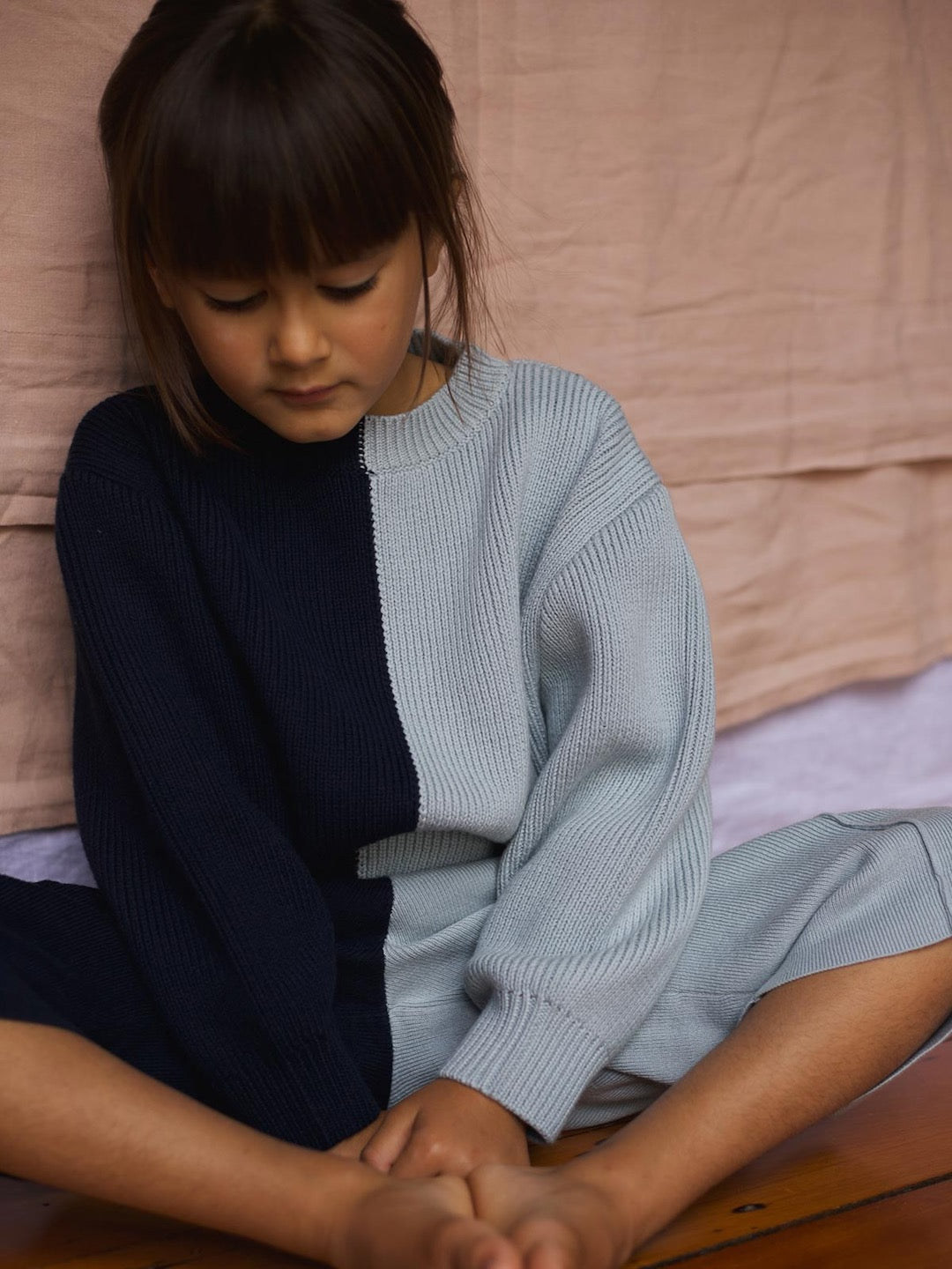 A little girl sitting on the floor in a Sol Sweater – Blue Multi by SUNNA STUDIOS.