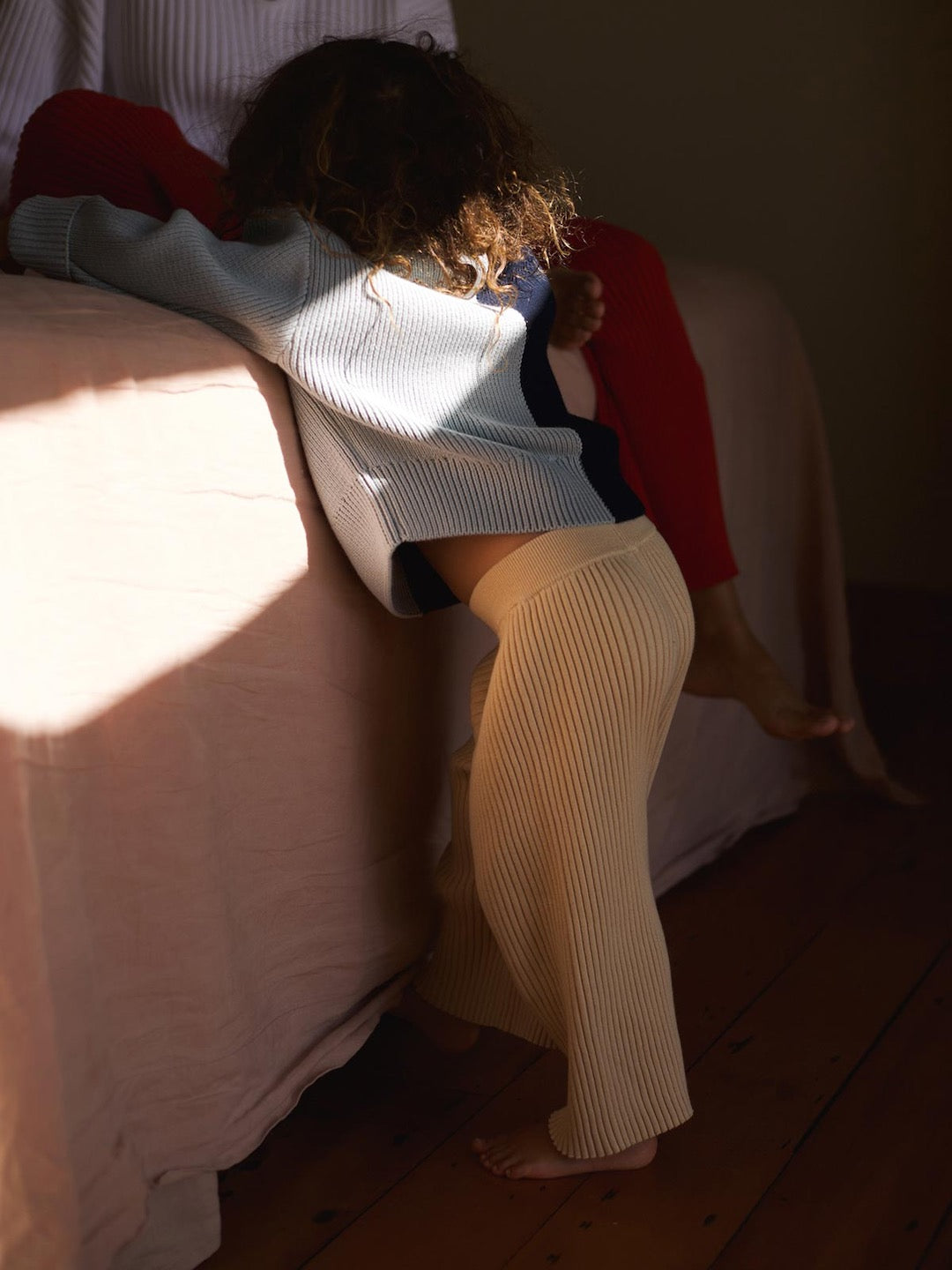 A little girl leaning on a bed wearing the Sol Sweater – Blue Multi by SUNNA STUDIOS.