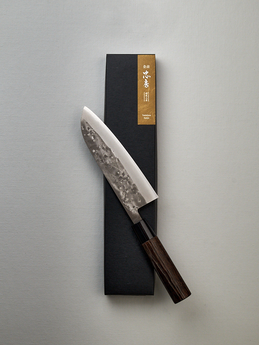 A Tadafusa Bocho Santoku Knife in a box on a white surface, suitable for all-purpose use with a focus on vegetables.