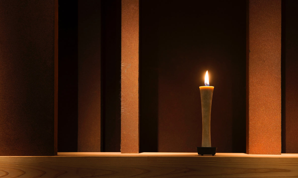 An IKARI Candle – Large (box of 1) sits on a wooden shelf in a dark room. Brand: Takazawa