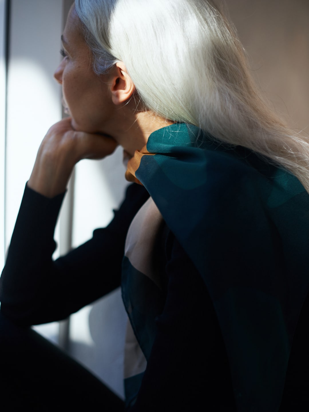 A woman with white hair looking out a window wearing a Poppy Scarf by Walker &amp; Bing.