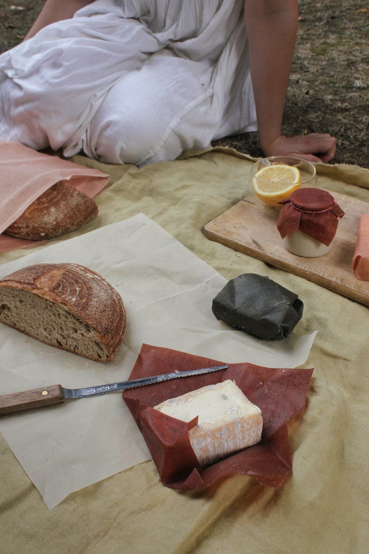 Person seated outdoors with a rustic picnic setup, featuring bread, cheese, and sliced citrus on a blanket, all neatly wrapped in Wax Atelier&#39;s Waxed Linen Food Wraps (Set of 3) – Ivy ⋄ Madder ⋄ Indigo instead of plastic clingfilm.