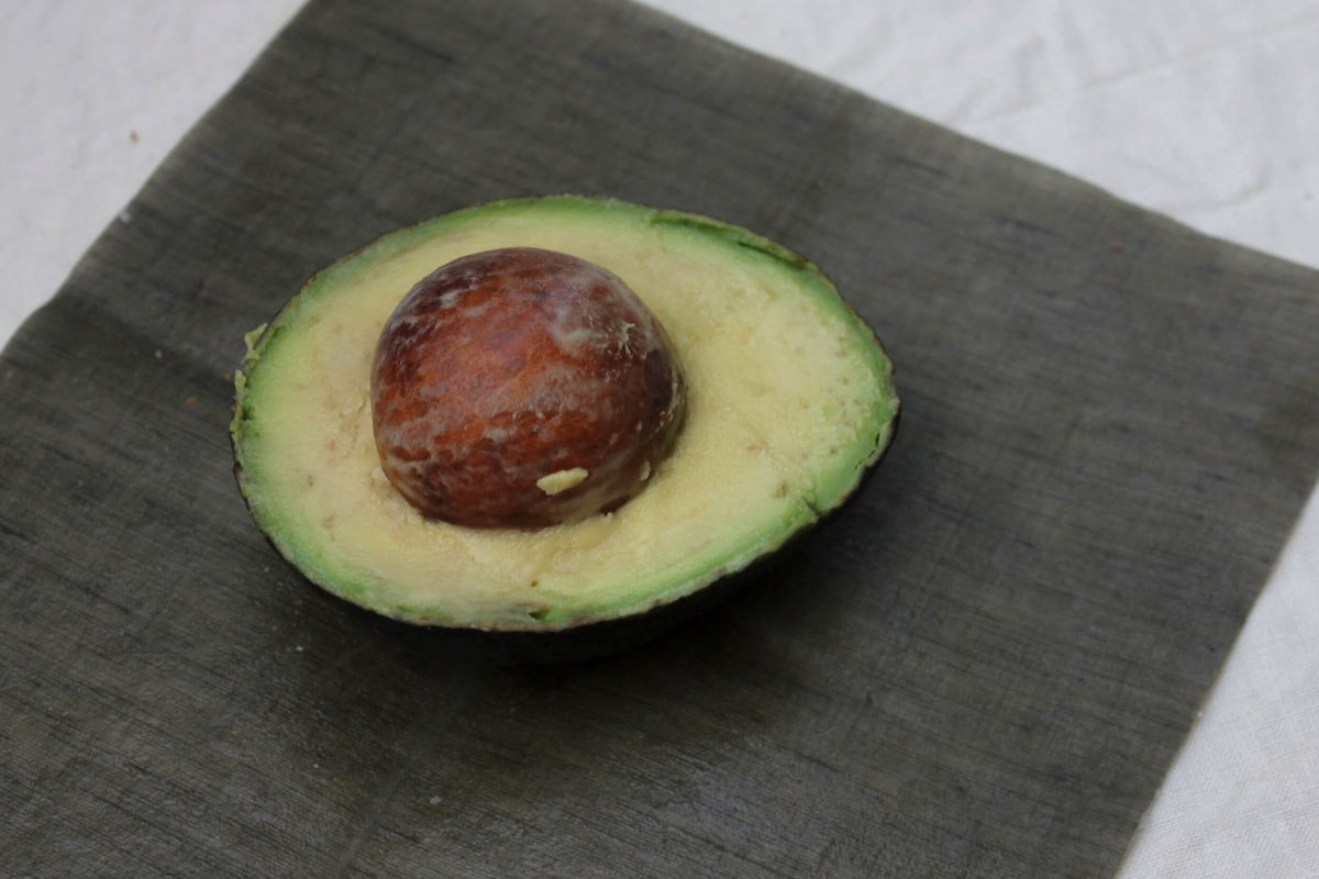 Half of an avocado with its pit on a wooden cutting board, now covered by Wax Atelier&#39;s Waxed Linen Food Wraps (Set of 3) – Ivy ⋄ Madder ⋄ Indigo as a sustainable alternative to plastic clingfilm.