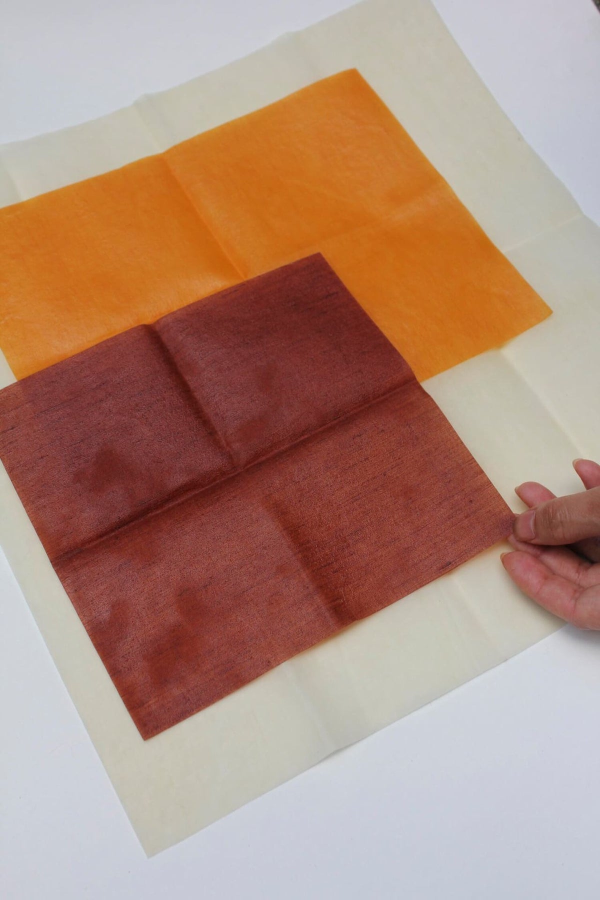 Hand positioning a piece of Ivy Waxed Linen Food Wraps fabric on top of an arrangement of orange dyed fabrics on a plain background, offering a sustainable alternative to plastic clingfilm. (Brand: Wax Atelier)