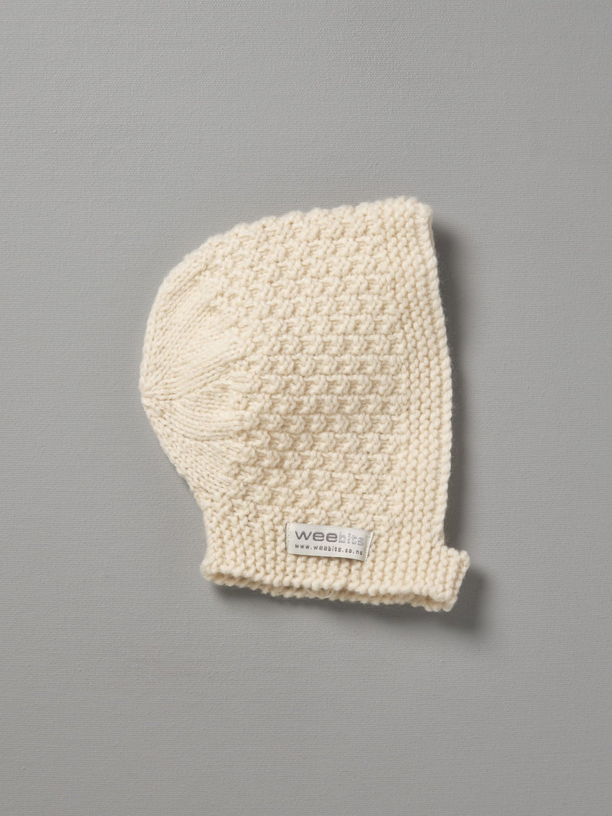 A Hand Knitted Baby Bonnet - Natural by Weebits on a grey background.