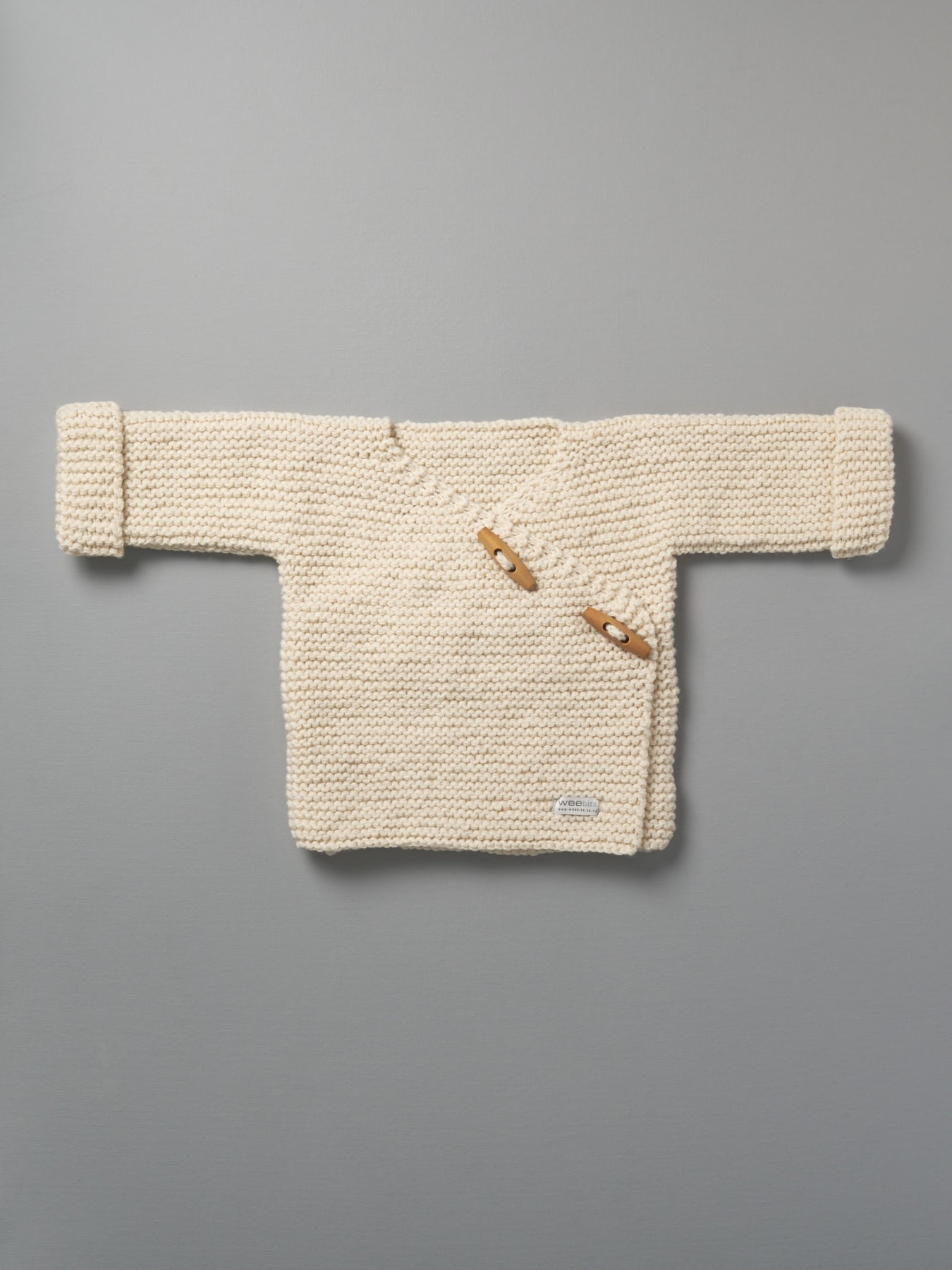 A Weebits Hand Knitted Double Breasted Jacket - Natural hanging on a gray background.