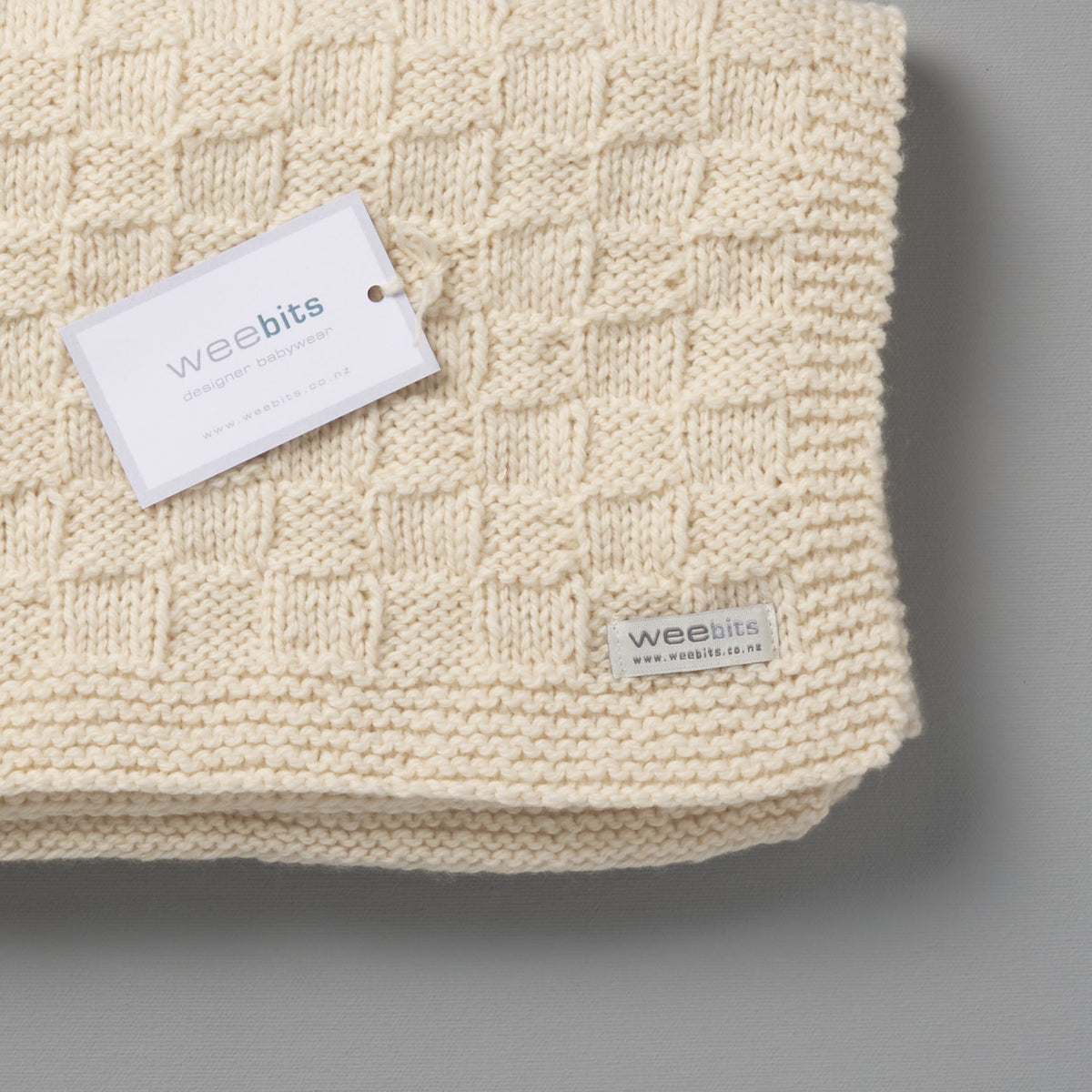 A white Hand Knitted Travel Rug - Natural with a tag on it, perfect for keeping babies warm and cozy. (Brand Name: Weebits)