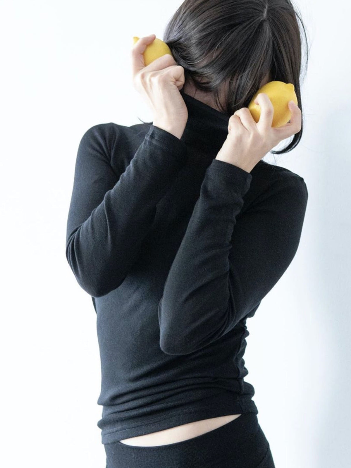 A woman wearing a YARN nz Tui Fine Merino Rib Turtle - Black and holding two lemons.