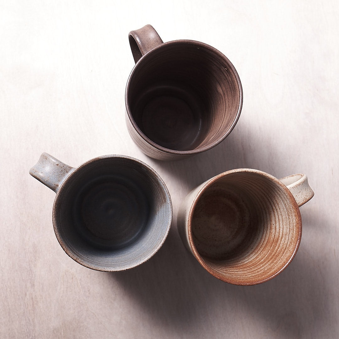 Three Zoë Isaacs grey-blue ceramic mugs on a wooden surface.
