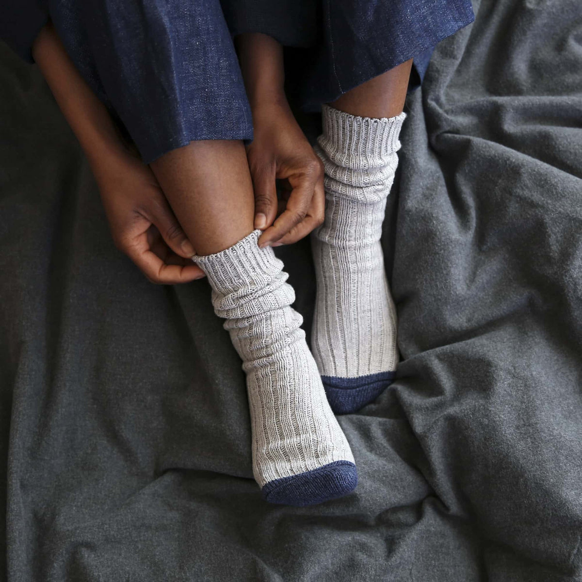 A woman wearing a pair of Boston Slab Socks - Grey Denim by Nishiguchi Kutsushita on a bed.