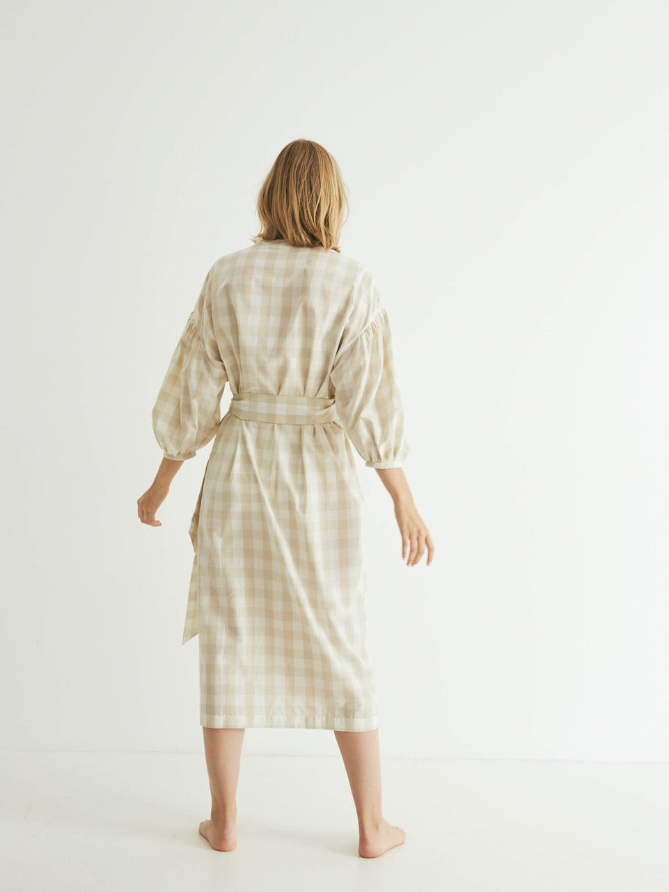 A person stands facing away from the camera, wearing a general sleep Agnes Wrap - Oatmeal Gingham with a tied belt.