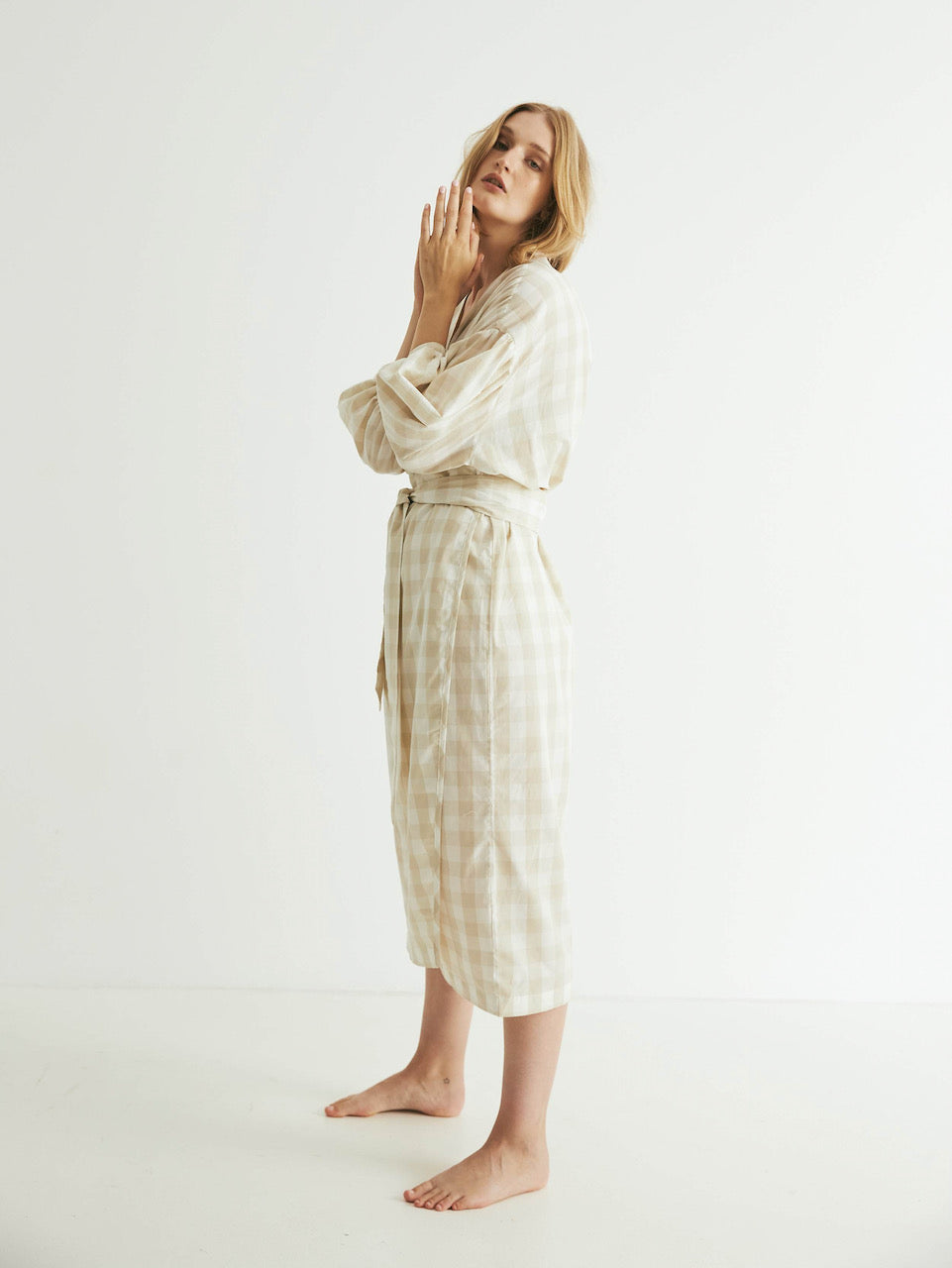 A person in a Agnes Wrap - Oatmeal Gingham robe stands barefoot with their hands gently touching their cheek and neck, epitomizing tranquility. (general sleep)
