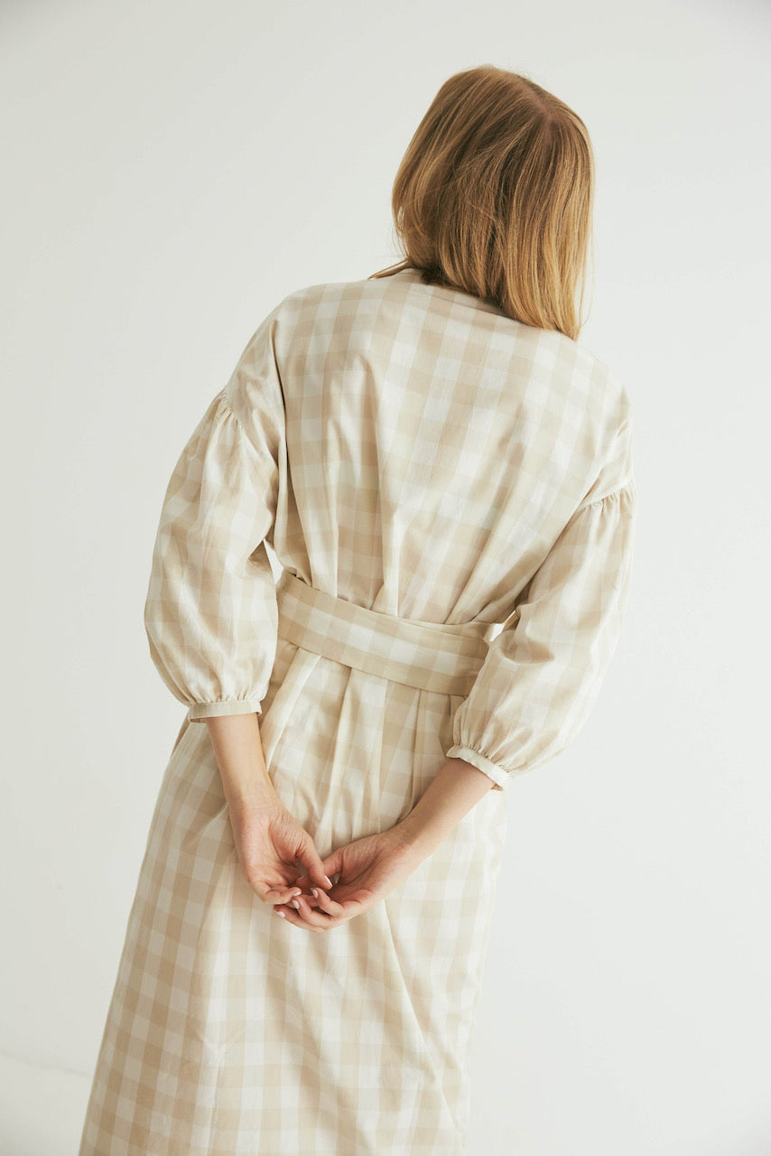 A woman standing with her back to the camera, wearing a long-sleeved Agnes Wrap - Oatmeal Gingham dress from general sleep and her hands clasped behind her back.